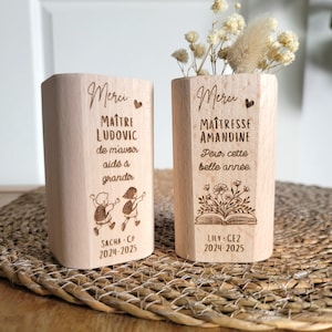 Peut inclure: Deux porte-crayons en bois avec texte et illustrations gravés. L'un dit "Merci Maître Ludovic" avec un dessin d'enfants et l'autre dit "Merci Maîtresse Amandine" avec un motif floral. Les deux ont des noms et des dates.