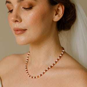 May include: A delicate necklace featuring alternating white pearls and amber-colored beads. The necklace is worn by a person with fair skin, wearing a white strapless dress, and a sheer veil. The person has their eyes closed and is wearing makeup.