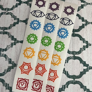 Può includere: Tre strisce di adesivi bianchi con simboli dei chakra in un arcobaleno di colori. Ogni striscia ha sette simboli, tra cui un fiore viola, un occhio, una stella blu, una stella verde, un triangolo arancione, un cerchio rosso e un quadrato rosso.