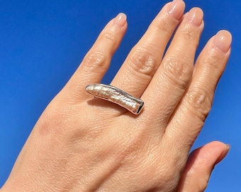 May include: A silver ring featuring a pearlescent, textured, rectangular-shaped stone. The ring is worn on a finger.