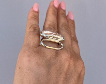 May include: A silver ring featuring an oblong top and a curved, open bottom band. The ring's top has a textured surface, and the design is modern and minimalist. The ring is worn on a hand with pink nail polish.