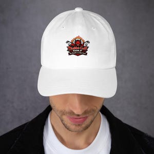 May include: A white baseball cap featuring a central graphic of a red devil character surrounded by orange flames, crossed golf clubs, and the text Diablo's Golf The Devil's in the Short Game printed in red, black, and white lettering on the front.