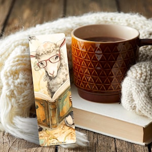 May include: A watercolor bookmark featuring a sheep wearing glasses and reading a book. The bookmark is next to a brown mug filled with a dark liquid, a white blanket, and a book. The text "LUNA AND LOOM" is at the bottom of the bookmark.