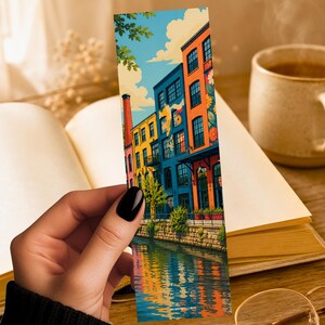 May include: A rectangular bookmark featuring a colorful illustration of multi-story buildings in shades of orange, yellow, and blue lining a canal. The scene includes a tall chimney, green trees, and a reflection of the buildings in the water below.
