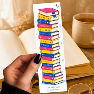 May include: A vertical rectangular bookmark featuring a colorful illustration of a tall stack of books in shades of pink, yellow, and blue. The books are decorated with small patterns like hearts and stars. The text reads: TBR taller than my trust issues.