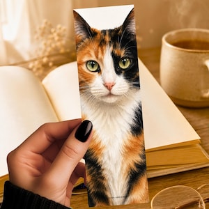May include: A rectangular bookmark featuring a detailed illustration of a calico cat with green eyes, white, orange, and black fur. The cat is shown from the chest up, facing forward. The text LUNA AND LEO is printed at the bottom of the bookmark.