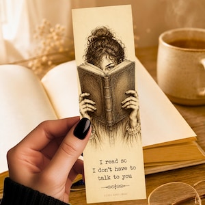 May include: A rectangular bookmark with a cream-colored background featuring a detailed black ink illustration of a person holding an open book in front of their face. The text reads, I read so I don't have to talk to you, followed by Luha and Lodac.