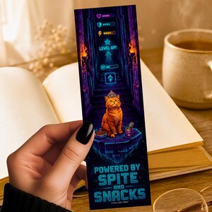 May include: A rectangular bookmark featuring a digital illustration of an orange cat wearing a crown, sitting on a stone platform. The background displays a neon-lit dungeon with game interface elements, including health and XP bars, and the text "LEVEL UP!" and "POWERED BY SPITE AND SNACKS".