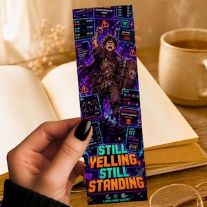 May include: A rectangular bookmark featuring a dark fantasy character illustration surrounded by digital game interface elements, status bars, and icons. The text "STILL YELLING, STILL STANDING" is printed in large, bold, cyan and orange letters at the bottom.