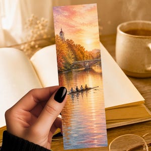 May include: A rectangular bookmark featuring a vertical illustration of a rowing team on a river at sunset. The scene includes a stone bridge, autumn trees, and a warm orange sky reflecting on the water. The text LUNA AND LOM is printed at the bottom.