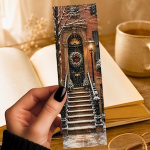 May include: A rectangular bookmark featuring a printed illustration of a brick building entrance with stone steps, a dark door with a green wreath, a glowing street lamp, and falling snow. The text LUNA AND LOOM is printed at the bottom.
