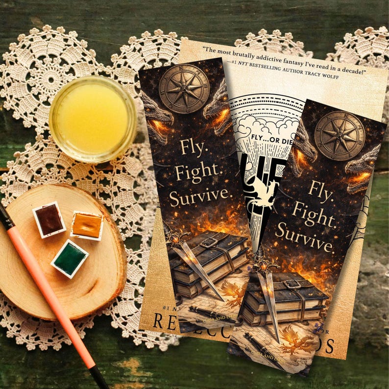 May include: Three fantasy-themed bookmarks with the text "Fly. Fight. Survive." feature dragon and compass designs. A glass of yellow liquid, watercolour paints, and a paintbrush are also visible.