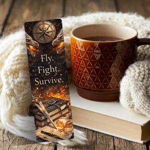 May include: A bookmark with the text "Fly. Fight. Survive." features a compass and dragon heads. A brown mug with a geometric pattern sits next to a book and a cosy white blanket. The scene evokes a fantasy theme.