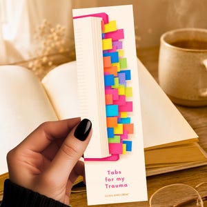 May include: A rectangular paper bookmark with a white background featuring a graphic of a white book spine with numerous colorful sticky tabs protruding from the pages. The text reads Tabs for my Trauma and LUNA AND LOOM in pink lettering.