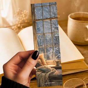 May include: A rectangular paper bookmark featuring a printed image of a window looking out onto a snowy scene. The design includes a cozy indoor setting with a knitted blanket, a small book, and a mug, with the text LUNA AND LOOM at the bottom.