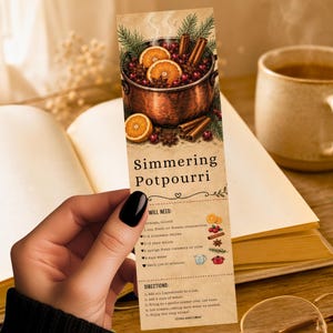 May include: A rectangular bookmark featuring a printed illustration of a copper pot filled with orange slices, cranberries, cinnamon sticks, and pine sprigs. The text reads Simmering Potpourri, followed by a list of ingredients and directions.