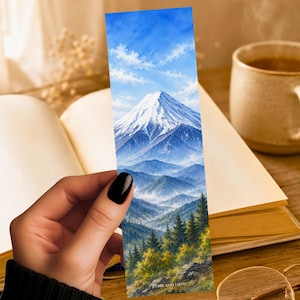 May include: A rectangular bookmark featuring a watercolor painting of a snow-capped mountain peak rising above layered blue hills and a dense forest of green and yellow trees under a bright blue sky with white clouds. The text FONA AND LOOM is visible.