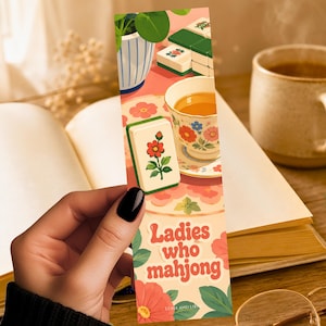 May include: A rectangular bookmark featuring a colorful illustration of mahjong tiles, a floral teacup, and a potted plant. The text "Ladies who mahjong" is printed in a bold, retro-style font at the bottom, surrounded by pink and red flower motifs.