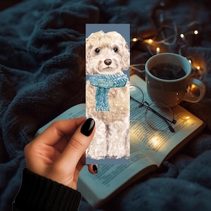 May include: A bookmark featuring a watercolor illustration of a fluffy, cream-colored dog wearing a blue scarf. The bookmark is held over an open book, with a cup of coffee and glasses in the background. The text "LUNA AND LOOSE" is printed on the bookmark.