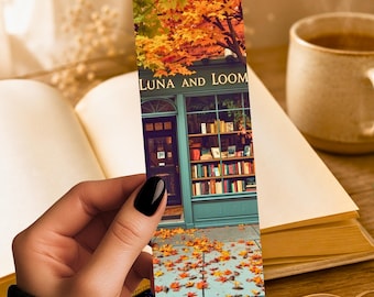 May include: A bookmark featuring a bookstore facade with the text "LUNA AND LOOM" and autumn foliage. The bookmark is held in a hand with black nail polish, with an open book and a cup of coffee in the background.