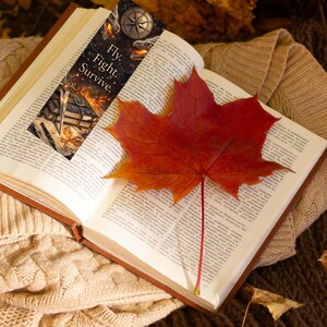 May include: An open book with a bookmark and a red maple leaf. The bookmark features the words "Fly. Fight. Survive." and a compass design. The leaf is a vibrant red and orange, and the book rests on a beige knitted sweater.