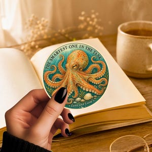 May include: A circular sticker featuring a detailed orange and yellow octopus illustration against a teal underwater background. The text reads THE SMARTEST ONE IN THE TANK and Luna and Loom. The design includes small seashells and aquatic plants.