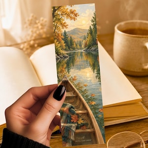 May include: A rectangular bookmark featuring a vertical illustration of a wooden canoe floating on a calm lake surrounded by autumn trees and mountains. A plaid blanket rests inside the canoe, and fallen orange leaves float on the water surface.