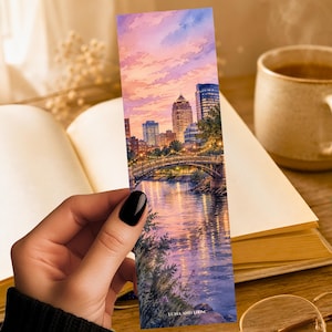 May include: A rectangular bookmark featuring a watercolor-style illustration of a city skyline at sunset over a river with an arched bridge. The scene includes purple and orange clouds, illuminated buildings, and the text LUNA AND LOOM at the bottom.