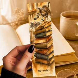 May include: A rectangular paper bookmark featuring a watercolor illustration of a tabby kitten chasing a small mouse atop a vertical stack of vintage books. The text Plot twist. and LUNA AND LOOM are printed in black at the bottom of the bookmark.