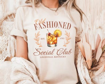 Puede incluir: Camiseta beige con el texto "Old Fashioned Social Club Louisville, Kentucky" en escritura marrón. El diseño incluye una ilustración de cóctel con una cereza y una rodaja de naranja, y el año 1783.