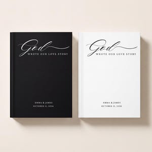 May include: Two hardcover books, one black and one white, each featuring the text God WROTE OUR LOVE STORY in elegant script and serif fonts, with the names EMMA & JAMES and the date OCTOBER 11, 2026 printed in a clean, minimalist sans-serif font.