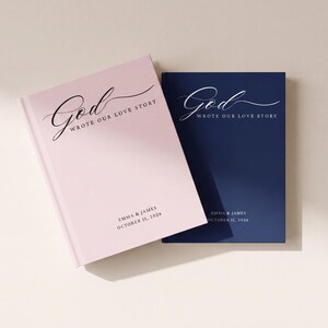 May include: Two hardcover books, one light pink and one navy blue, each featuring the white text God WROTE OUR LOVE STORY in a script and serif font, with the names EMMA & JAMES and the date OCTOBER 11, 2026 printed in a simple sans-serif font.