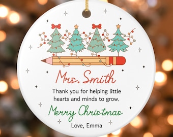 May include: A white ceramic Christmas ornament with a gold string. The ornament features four green Christmas trees with red bows and string lights atop a yellow pencil. Text includes "Mrs. Smith" and "Merry Christmas."
