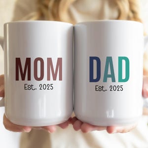 May include: Two white ceramic mugs with the words "MOM Est. 2025" and "DAD Est. 2025" in colorful block letters.