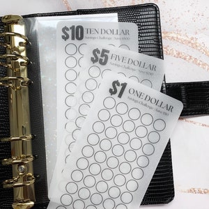 May include: Three clear plastic sheets with circles for tracking savings. The sheets are labelled "$10 Ten Dollar Savings Challenge Save $500", "$5 Five Dollar Savings Challenge Save $250", and "$1 One Dollar Savings Challenge Save $50". The sheets are in a black binder.