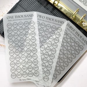 May include: Translucent savings challenge cards with black printed dollar amounts. The cards are labelled "One Thousand", "Two Thousand", and "Three Thousand Dollar Savings Challenge". The cards are in a black binder with gold rings.