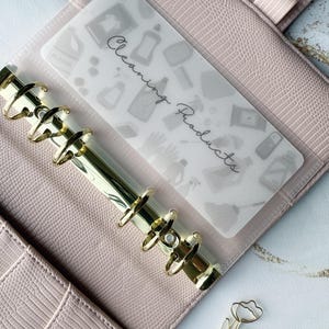 May include: A light pink planner with gold rings and a clear insert labeled "Cleaning Products." The insert features a pattern of cleaning supplies. A gold tulip paperclip rests on the surface.