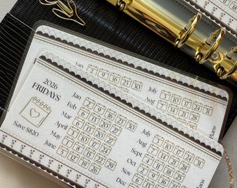 May include: A black leather planner with gold-colored rings and a gold-colored tulip paper clip. The planner includes calendar inserts for 2026, with dates and the text "FRIDAYS" and "Save $820".