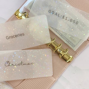 May include: A light pink, textured binder with gold rings holds several clear, sparkly envelopes. The envelopes are labeled "Groceries", "Christmas", and "GOAL: $1,000". The envelopes contain cash, and the binder is open.