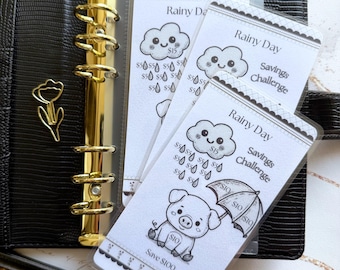 May include: A black planner with gold accents and a tulip-shaped paper clip. Several white cards with a cloud and pig illustration are visible. The cards have text that reads "Rainy Day Savings Challenge."
