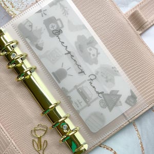 May include: A light pink planner with gold accents, featuring a clear insert labeled "Emergency Fund." The insert is decorated with small, gray illustrations. A gold tulip-shaped paper clip is visible.