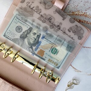 May include: A pink faux leather binder with gold hardware, holding a transparent sleeve labelled "Vehicle Maintenance." The sleeve contains a &pound;79.00 bill. A gold tulip-shaped paper clip rests on the surface.