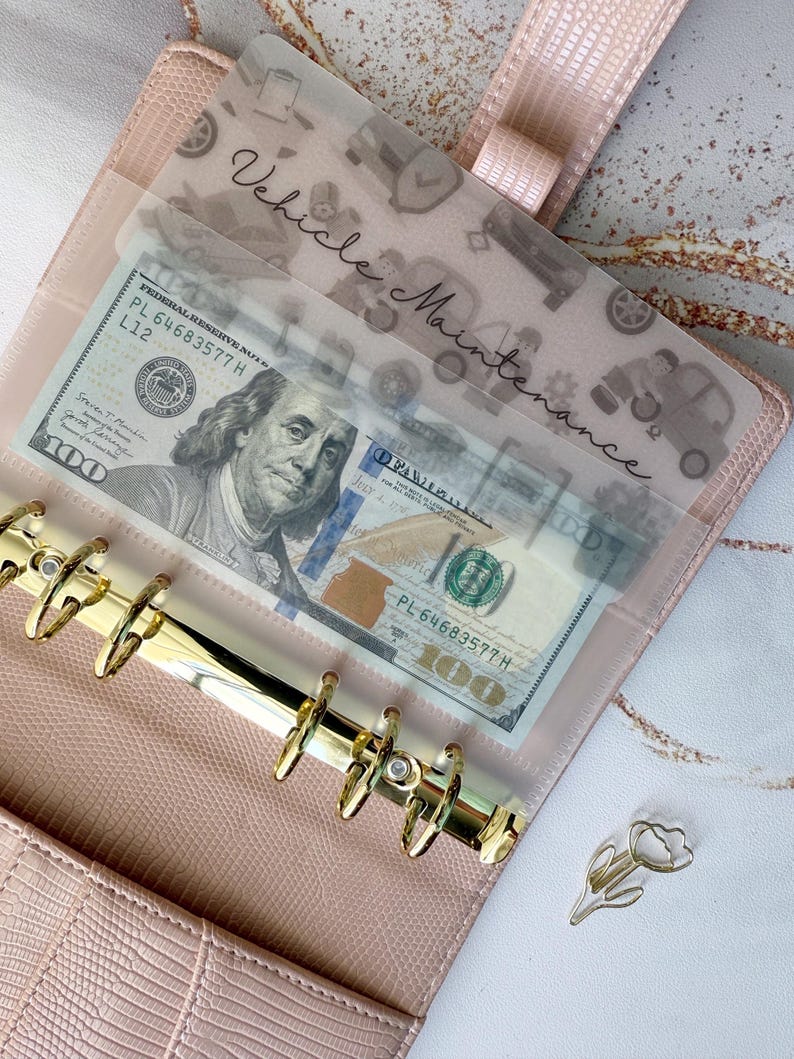 May include: A pink faux leather binder with gold hardware, holding a transparent sleeve labelled "Vehicle Maintenance." The sleeve contains a &pound;79.00 bill. A gold tulip-shaped paper clip rests on the surface.