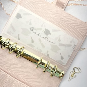 May include: A light pink planner with gold accents, featuring a clear insert with graduation-themed graphics and the word "Graduation." A gold tulip-shaped paper clip rests beside the planner. The planner has a textured, faux-leather appearance.