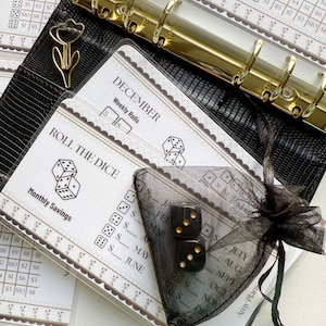May include: A black faux leather binder with gold rings, a gold flower paper clip, and inserts for monthly savings. Two black dice with gold dots are in a sheer black bag. The inserts have text that reads "Roll the Dice" and "December Weekly Rolls."