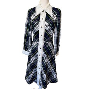 May include: A long-sleeved knee-length dress featuring a dark blue, gray, and white plaid pattern with thin yellow accent lines. The dress has a solid white pointed collar, white cuffs, and a central vertical white button placket.