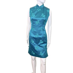 May include: Teal sleeveless dress with a mandarin collar and floral embroidery. The dress features a fitted silhouette and decorative buttons on the sides. The fabric has a subtle sheen, adding to its elegant appearance. A classic, stylish garment.