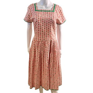 May include: A knee-length dress featuring a cream base with a repeating red and green floral pattern. The garment has short sleeves, a square neckline trimmed in green, and a gathered waistline that flows into a pleated skirt with a green hem.