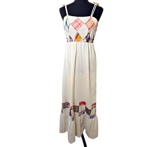 May include: A cream-colored maxi dress with a patchwork bodice and hem. The bodice features diamond-shaped patches in various plaid patterns. The dress has thin white straps and a ruffled hemline, creating a vintage, bohemian style.