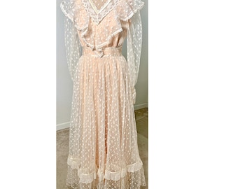 May include: A vintage-style, floor-length dress in a soft peach color. The dress features a high, lace-trimmed collar, ruffled bodice, and sheer, dotted sleeves. The skirt has a layered design with a ruffled hem, creating a romantic and elegant look.